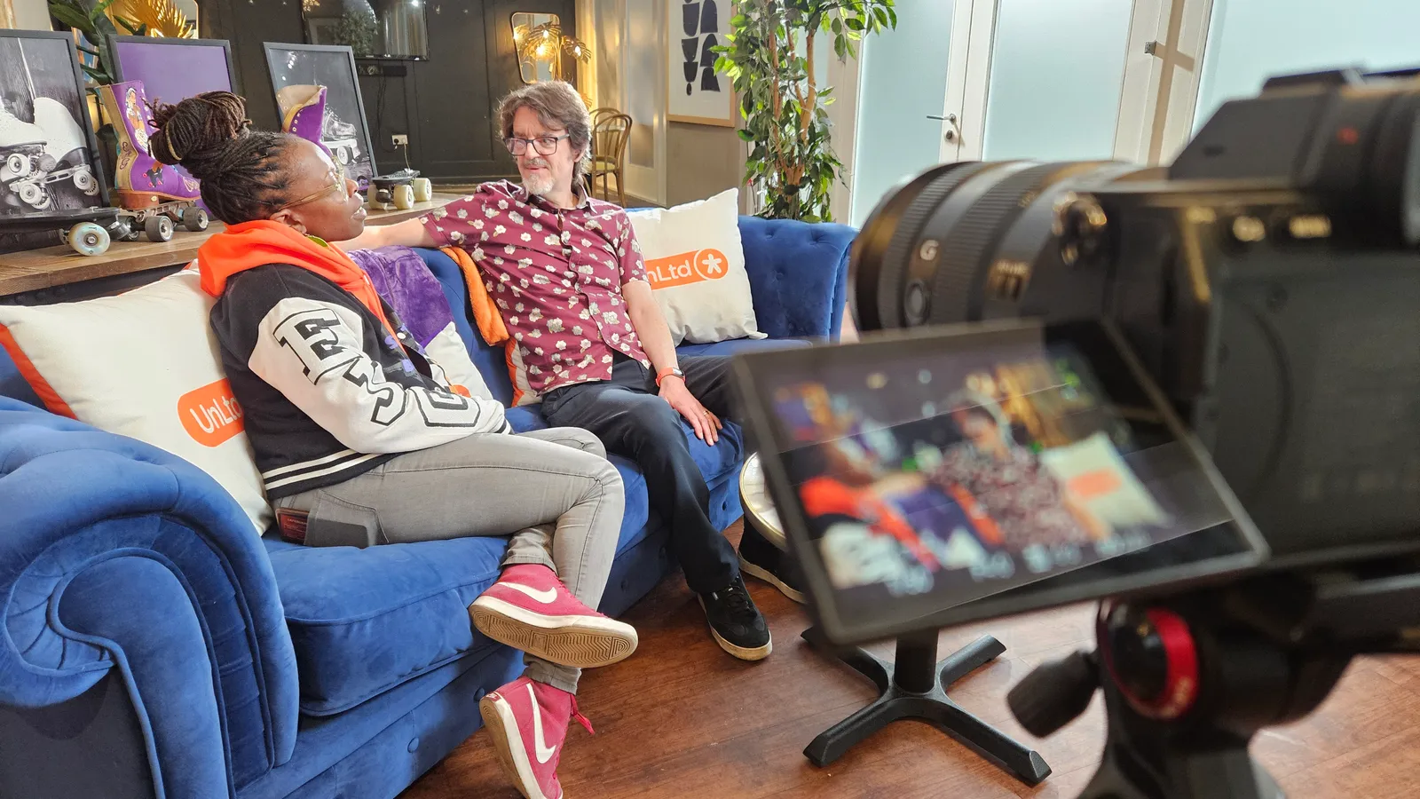 Empress and John sit on a blue sofa together while we look through a camera that is filming them.