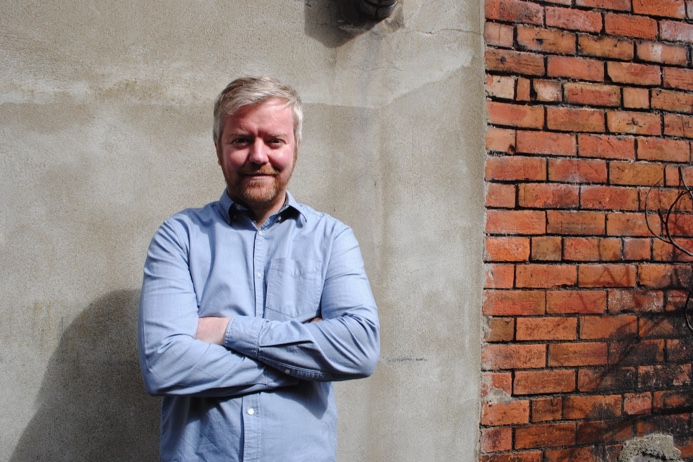 Rory stands against a brick wall, facing the camera with his arms folded. He has a ginger beard, grey swept hair and is wearing a blue shirt.