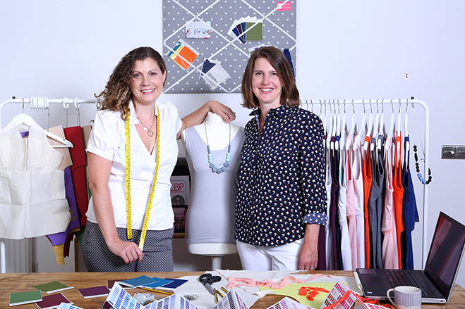 Philippa and Lisa stand either side of a dress makers dummy, in front of a rack of clothes, with fabric cutting scraps on a table in front of them.